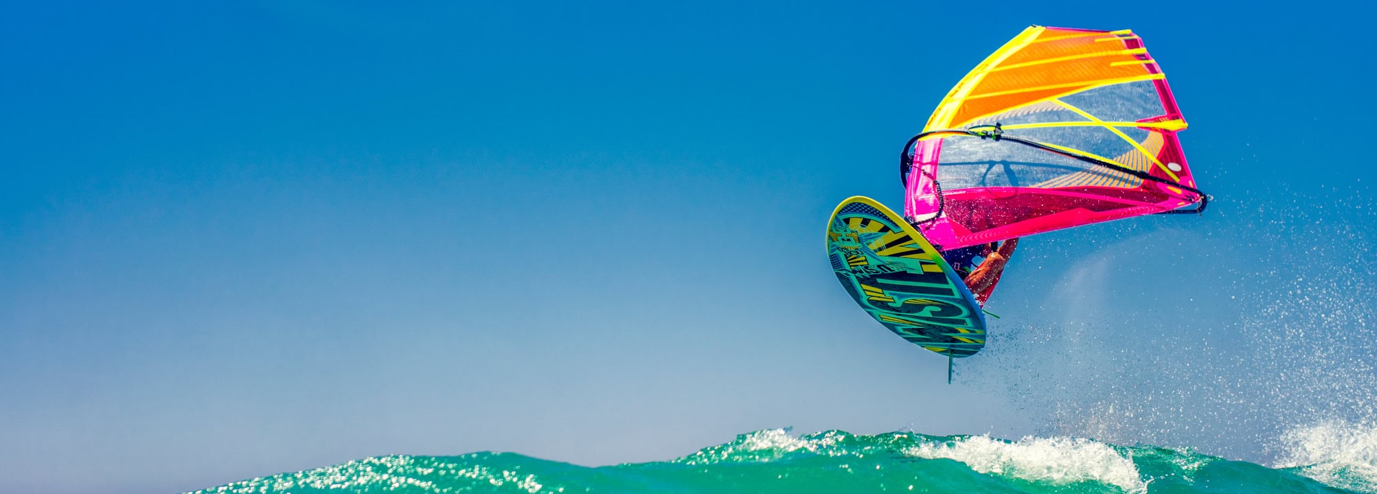 Bright pink and yellow kite surfing sail soaring above turquoise ocean waves against a clear blue sky.